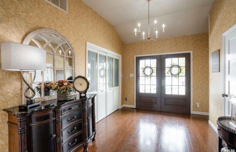 Bright foyer with chandelier, decorative mirror, and wooden doors with wreaths. Elegant entryway design.