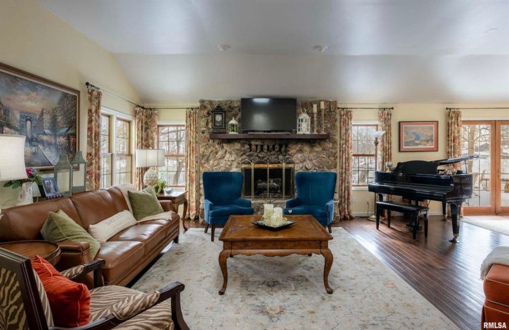 Cozy living room with stone fireplace, blue armchairs, and grand piano, featuring warm decor.
