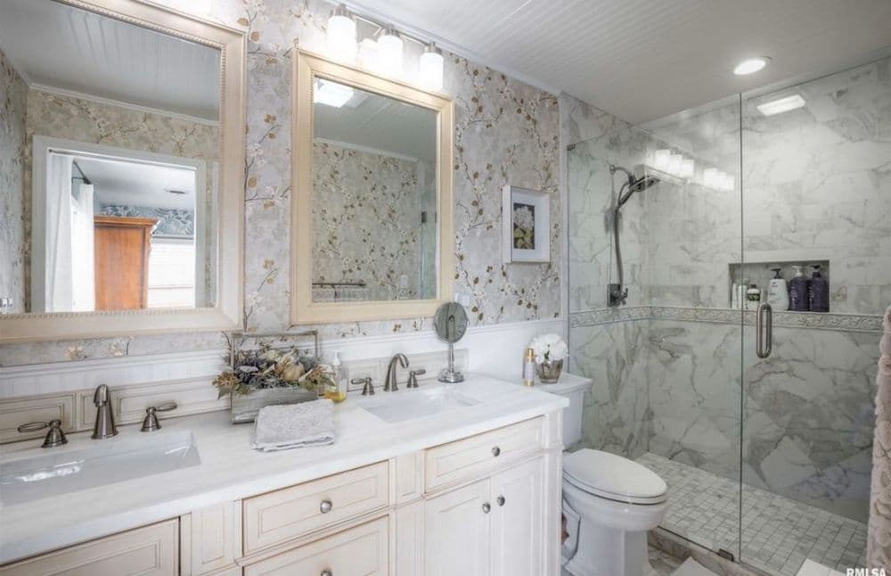 Modern bathroom with double sink, glass shower, marble tiles, and floral wallpaper accents.