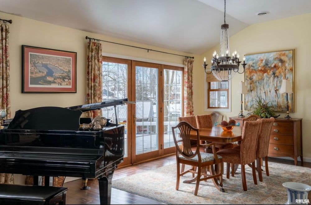Elegant dining room with a grand piano, chandelier, and wooden dining table by large windows.
