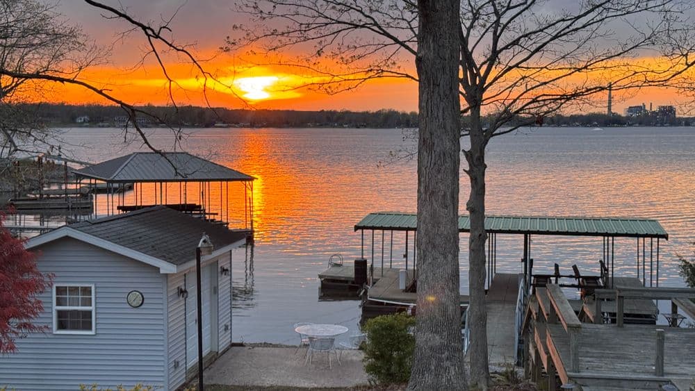 Sunset over calm lake with docks and a cabin, reflecting vibrant orange and yellow hues.