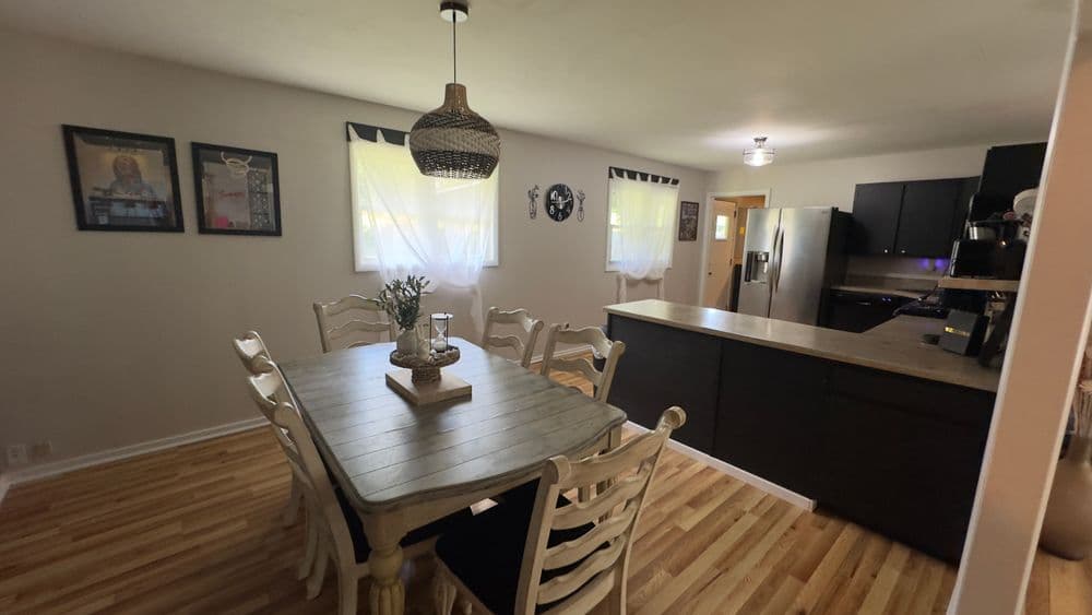 Bright dining room with wooden table, black kitchen cabinets, and natural light from windows.