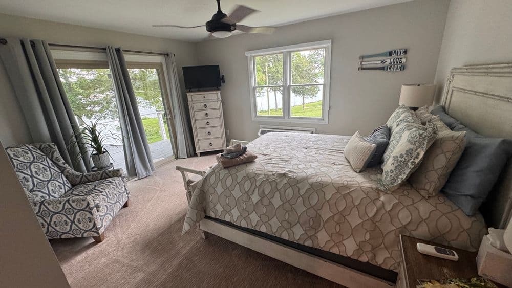 Cozy bedroom with lake view, featuring a queen bed, armchair, and TV in natural light.