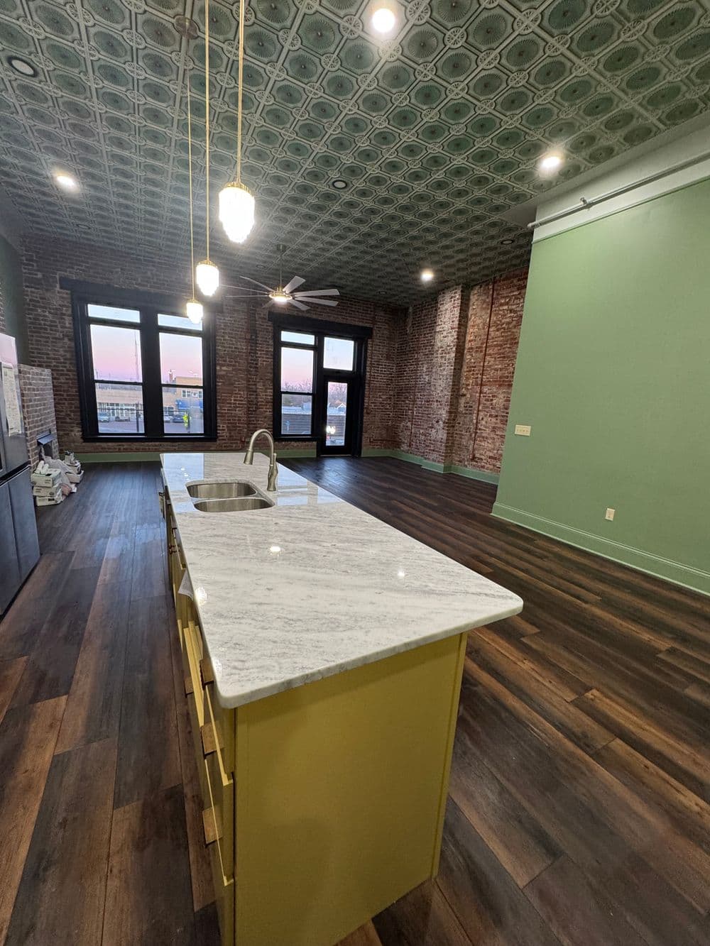 Modern kitchen with a marble island, exposed brick walls, and decorative ceiling tiles at sunset.