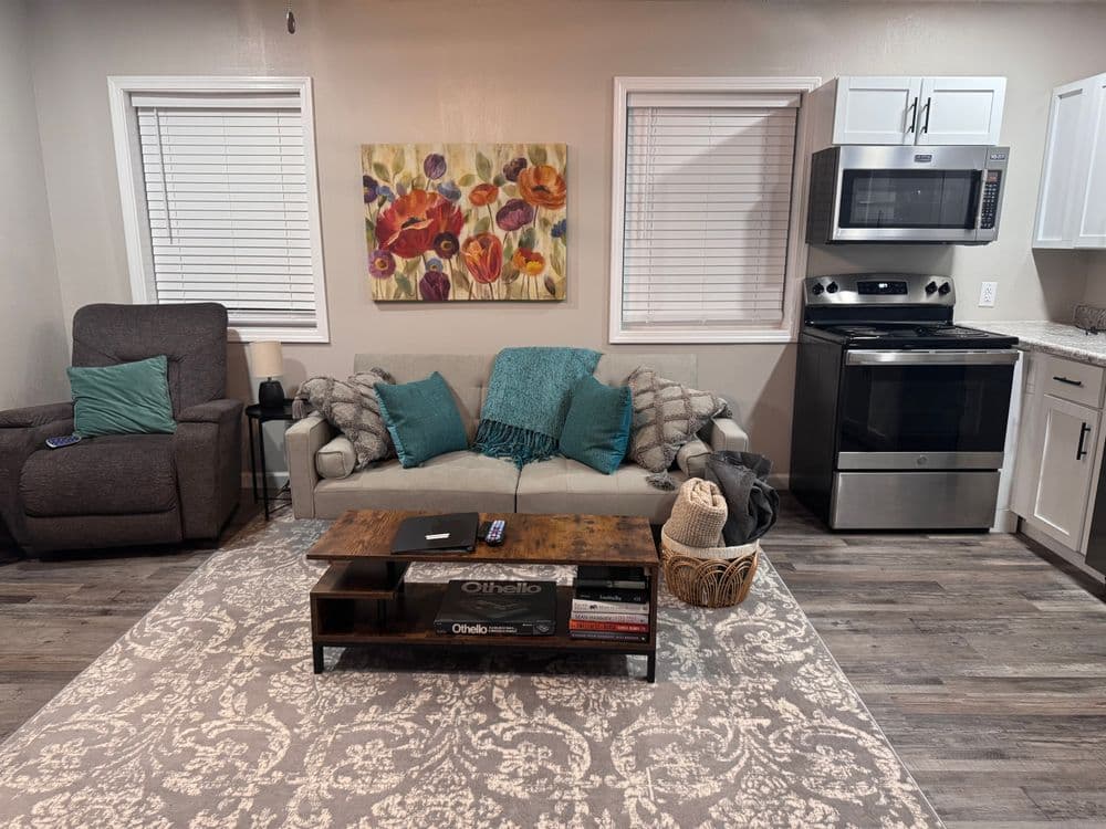 Cozy living room with couch, floral art, gray armchair, and modern kitchen appliances.